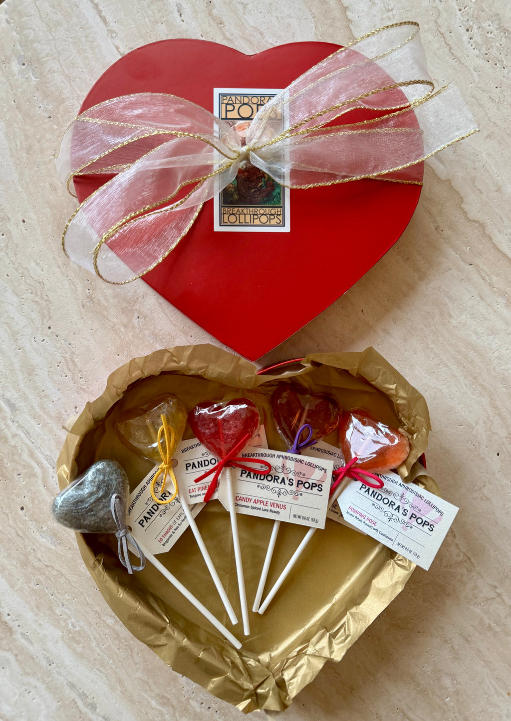 A heart-shaped red box with a clear bow containing various flavored lollipops displayed in a brown paper dish. Each lollipop has a label indicating its flavor.
