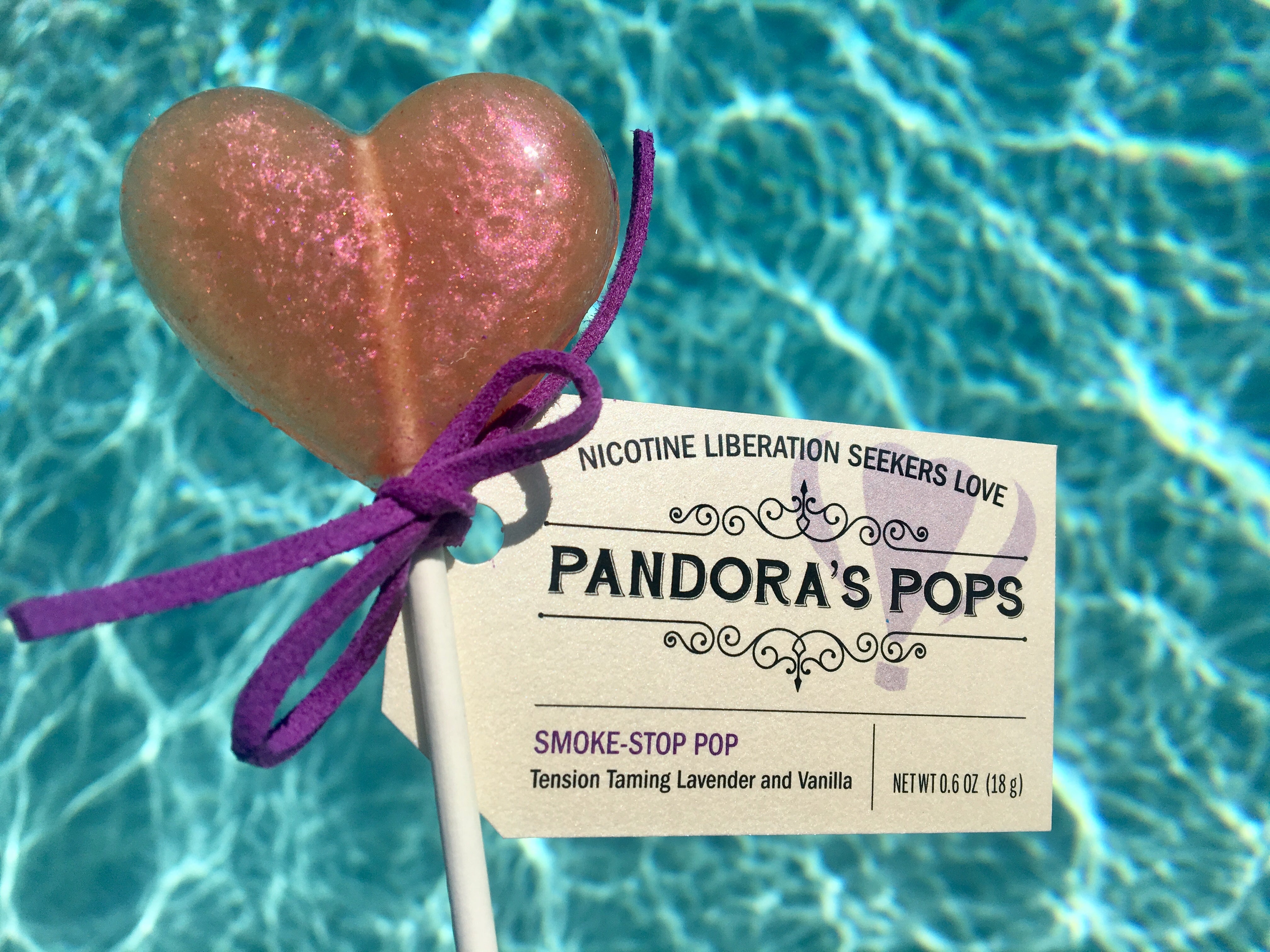 A heart-shaped lollipop with a pink candy coating, on a stick, with a purple ribbon label that reads 'Pandora's Pops - Smoke Stop Pop - Tension Taming Pop: Lavender and Vanilla'.